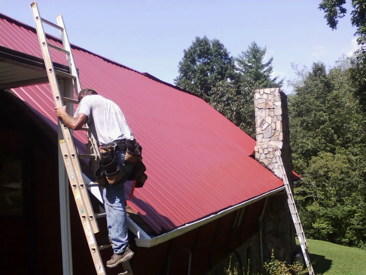 Roofer on a ladder beside a red metal roof and stone chimney — Soffit & Fascia Repair by Roofing Brothers in Whiteland